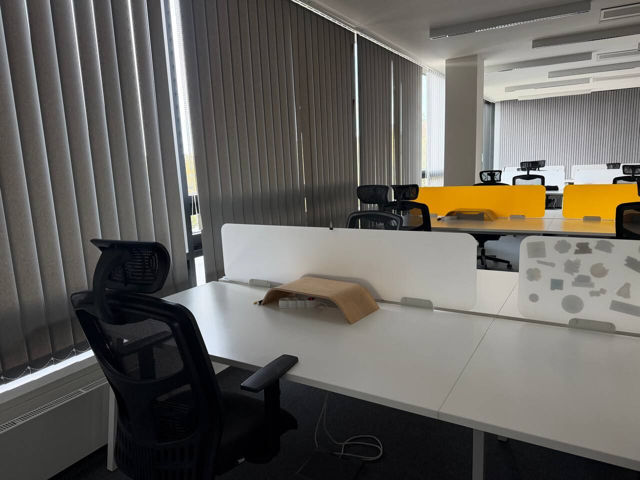 Open space with rows of desks, black chairs, yellow desk dividers and vertical blinds